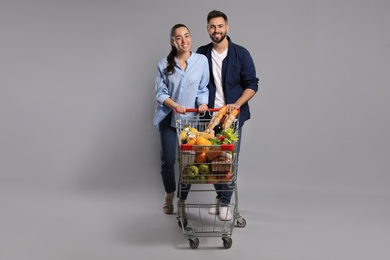 Happy couple with shopping cart full of groceries on light grey background
