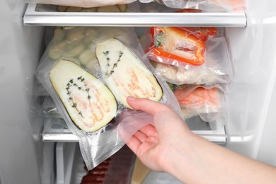 Photo of Woman putting vacuum bag with eggplant into fridge, closeup. Food storage