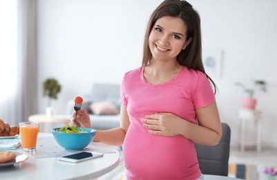 Young pregnant woman eating vegetable salad at table in kitchen