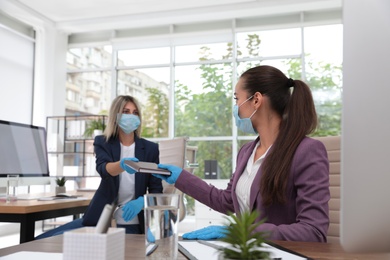 Office employee in mask and gloves giving notebook to her colleague at workplace