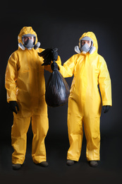 Man and woman in chemical protective suits holding trash bag on black background. Virus research
