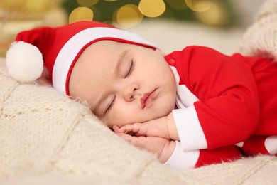 Cute baby in Christmas costume sleeping on knitted blanket against blurred festive lights. Winter holiday