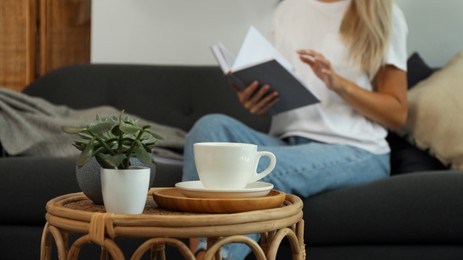 Photo of Woman reading book indoors, focus on table with cup and houseplants