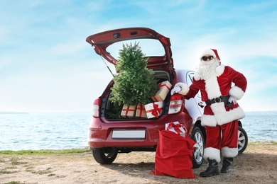 Photo of Authentic Santa Claus near red car with gift boxes and Christmas tree on beach