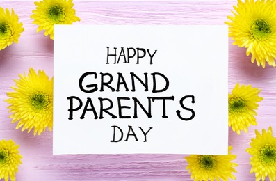 Beautiful yellow flowers and card with phrase Happy Grandparents Day on pink wooden background, flat lay