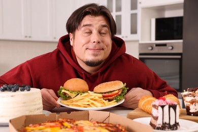 Happy overweight man holding plate with tasty burgers and French fries at table in kitchen