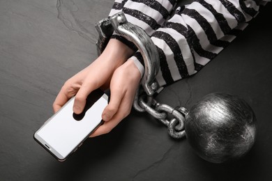 Prisoner shackled with ball and chain holding smartphone at black table, top view. Internet addiction
