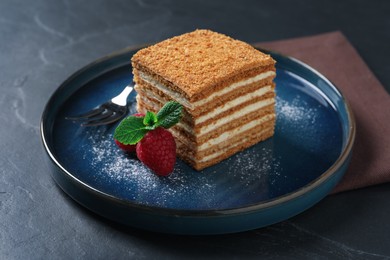 Slice of delicious layered honey cake with mint, raspberries and powdered sugar served on black table
