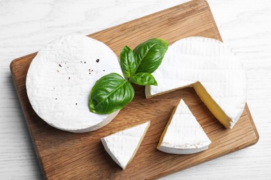 Tasty cut and whole brie cheeses with basil on white wooden table, top view