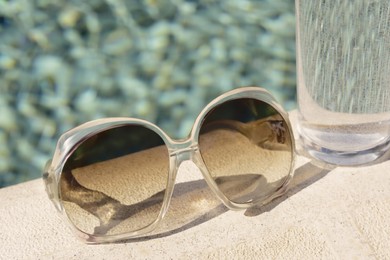Photo of Stylish sunglasses and glass of water near outdoor swimming pool on sunny day, closeup