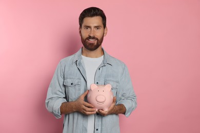 Happy man with ceramic piggy bank on pale pink background