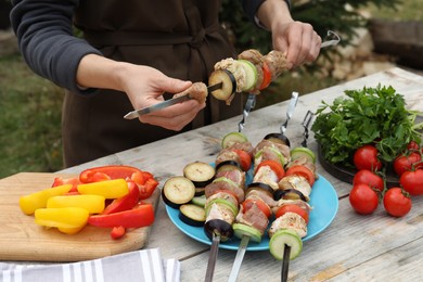 Woman stringing marinated meat and vegetables on skewer at wooden table outdoors, closeup