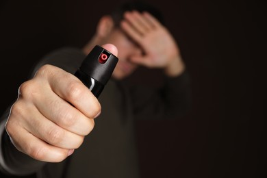Man covering eyes with hand and using pepper spray on black background, focus on canister