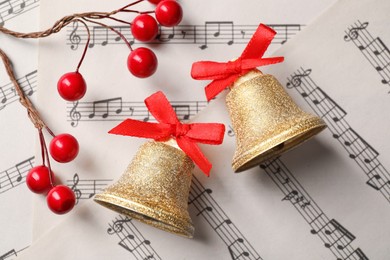Photo of Golden shiny bells with red bows and decorative berries on music sheets, flat lay. Christmas decoration