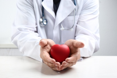 Doctor with red heart at white table indoors, closeup