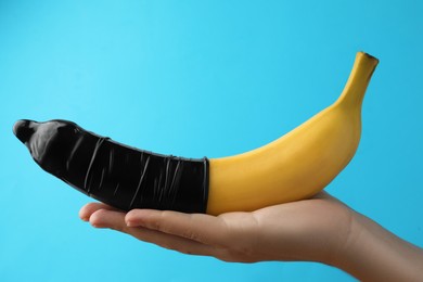 Woman holding banana in condom on light blue background, closeup. Safe sex concept