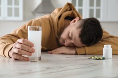 Man sleeping at table in kitchen, focus on hand with glass of medicine for hangover