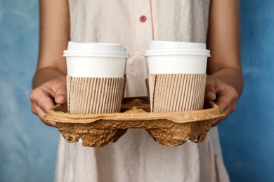 Woman holding cardboard holder with takeaway paper coffee cups on light blue background, closeup