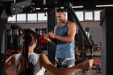 Photo of Happy trainer writing down plan of workouts while woman doing exercise in modern gym