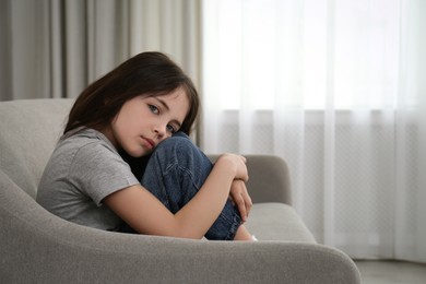 Sad little girl sitting on sofa indoors, space for text