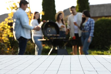 Empty white wooden table and blurred view of friends having barbecue party outdoors. Space for text