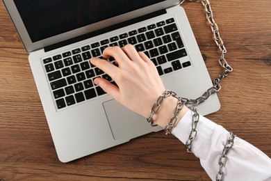 Photo of Woman with chained hand typing on laptop at wooden table, top view. Internet addiction