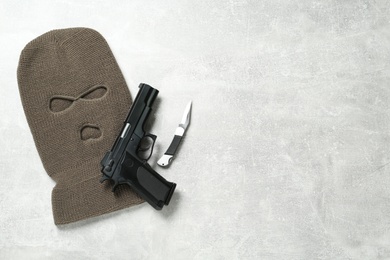 Beige knitted balaclava, pistol and knife on grey table, flat lay. Space for text