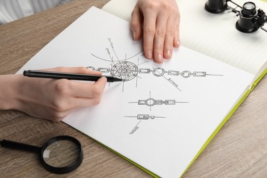 Photo of Jeweler drawing sketch of elegant bracelet on paper at wooden table, closeup