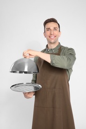Handsome waiter holding metal tray with lid on light background