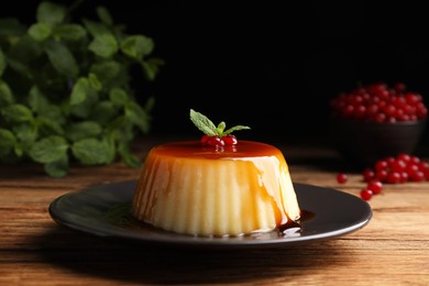 Photo of Plate of delicious caramel pudding with red currants and mint on wooden table against black background