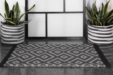 Stylish door mat with beautiful pattern and green houseplants on floor