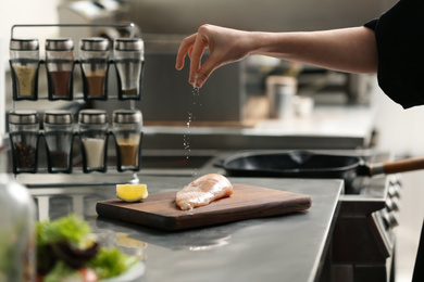 Female chef adding salt to raw chicken fillet in restaurant kitchen, closeup. Cooking food