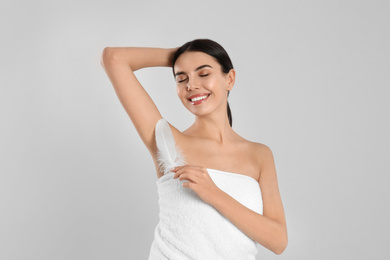 Young woman touching armpit with feather after epilation procedure on light grey background