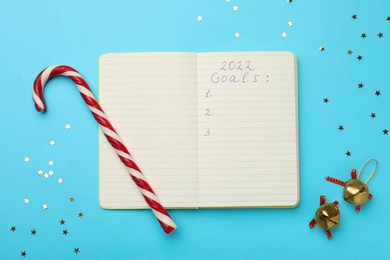 Open planner, confetti, candy cane and bells on light blue background, flat lay. Planning for 2022 New Year