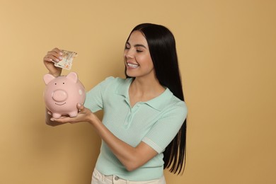 Happy young woman putting money into piggy bank on beige background