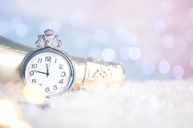 Pocket watch and bottle of champagne on snow against blurred lights, space for text. New Year countdown