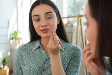 Photo of Beautiful young woman applying cosmetic pencil on lips near mirror indoors