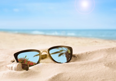 Green palm leaves mirroring in sunglasses on sandy beach with seashell near sea