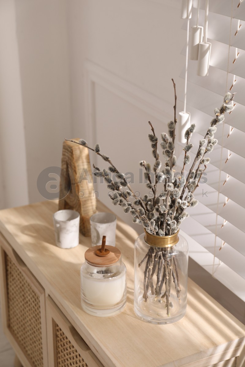Glass vase with pussy willow tree branches and candle on wooden commode near window in room