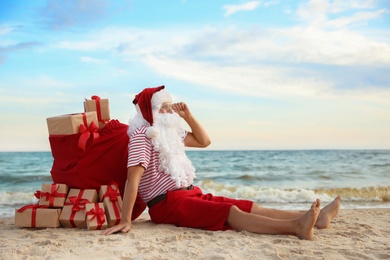 Photo of Santa Claus with bag of presents relaxing on beach, space for text. Christmas vacation