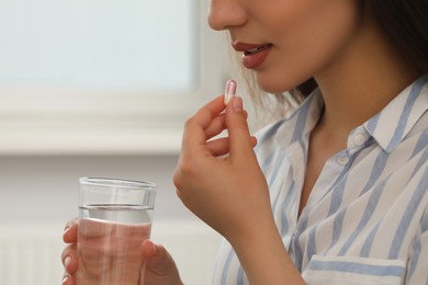 Young woman with glass of water taking dietary supplement pill indoors, closeup