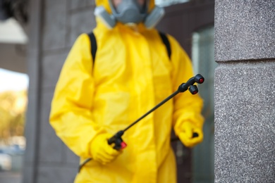 Person in hazmat suit disinfecting building wall outdoors, focus on sprayer. Surface treatment during coronavirus pandemic