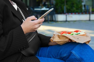 Woman holding tasty sandwich with vegetables and using phone outdoors, closeup. Street food