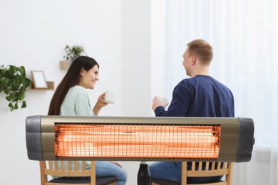 Photo of Couple spending time in room with modern electric infrared heater, focus on appliance