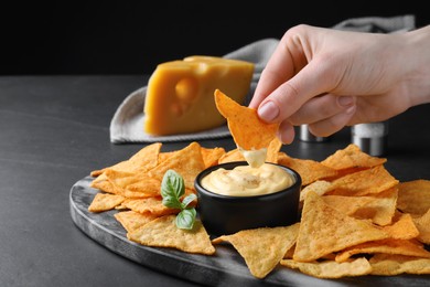 Woman dipping crispy nacho into delicious cheese sauce at black table, closeup. Space for text