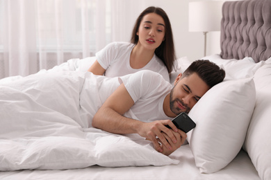 Distrustful young woman peering into boyfriend's smartphone in bed at home