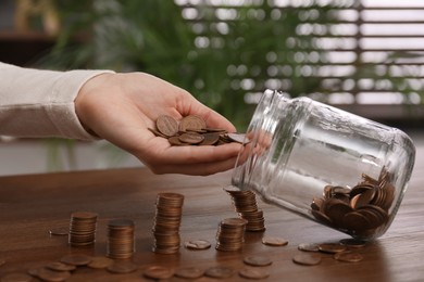 Woman putting coins into glass jar at wooden table indoors, closeup