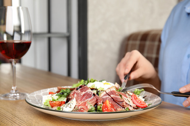 Woman eating delicious prosciutto salad at wooden table indoors, closeup. Space for text