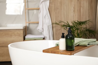 Wooden bath tray with bottles of shower gels and towel on tub indoors, space for text