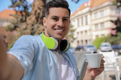 Happy man with cup of coffee and headphones taking selfie in outdoor cafe
